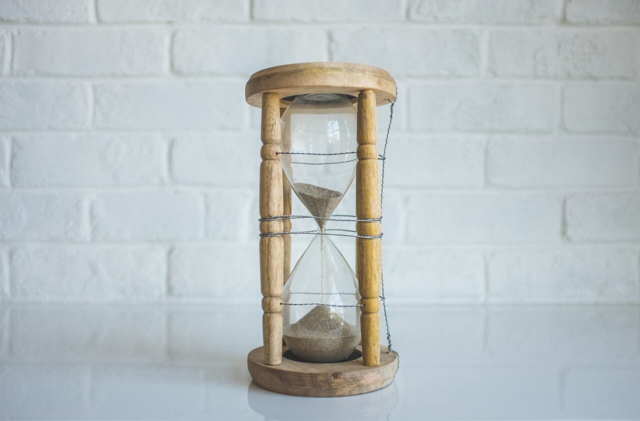 a wooden hourglass on a white tabletop in front of a white brick wall.