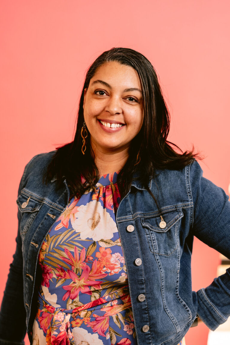 A mixed race woman with shoulder length dark hair in front of a salmon colored background. She is wearing a floral jumpsuit and jean jacket.