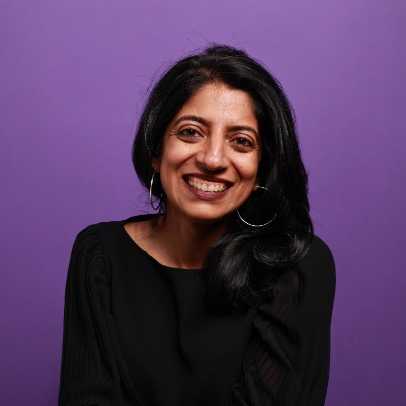 A bust-style headshot of a south Asian woman with long dark hair against a purple background.