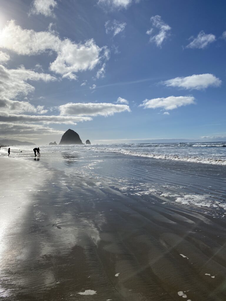 The beach in the foreground fills half the image, and the blue sky with white puffy clouds fill the other half. Haystack Rocks in Cannon Beach, OR dot the horizon.