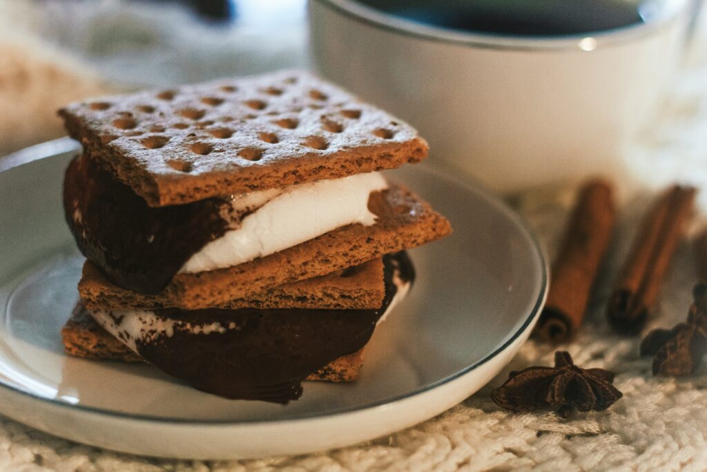 A double-stacked s'mores on a plate. Two cinnamon sticks and a mug are next to it.